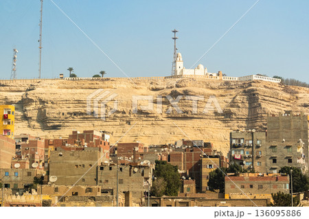A white mosque stands atop a cliff in Cairo, overlooking a densely populated area. The mosque features a tall minaret and is surrounded by rocky terrain. 136095886