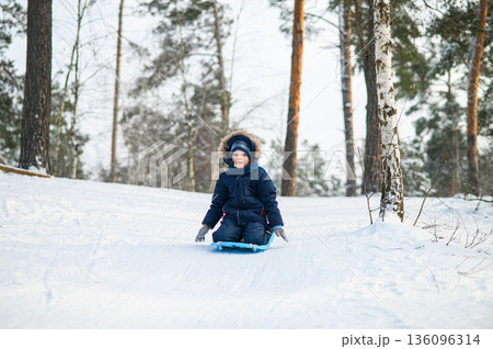 Little boy enjoying winter outdoor fun while sledding in snowy forest. Active childhood, seasonal outdoor recreation, winter adventure, family leisure and healthy lifestyle concept 136096314