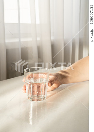 Hand picking up a glass of transparent water on a white table, promoting hydration, health, and wellness Hand picking up a glass of transparent water on a white table, promoting hydration, health, and wellness 136097153