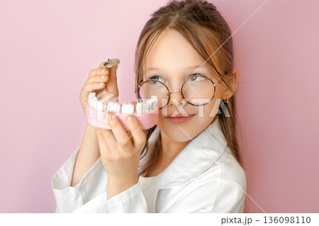 Child dressed as dentist holding teeth model on pink background. Concept of World Oral Health Day, oral hygiene awareness, prevention, healthy teeth education and children dental healthcare. Child dressed as dentist holding teeth model on pink background. Concept of World Oral Health Day, oral hygiene awareness, prevention, healthy teeth education and children dental healthcare. 136098110