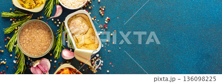 A top view of open tin cans with canned tuna, mackerel, shrimp, sardine, tuna, Cod liver, surrounded by spices and herbs on a blue background, banner 136098225