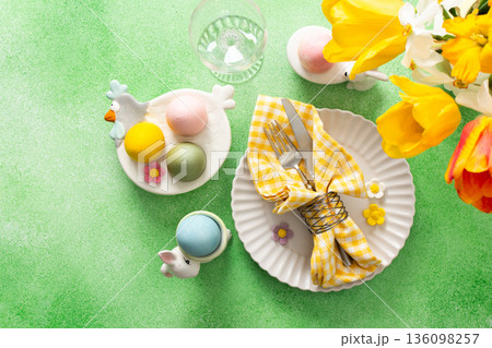 A beautifully arranged Easter table setting showcases a decorative egg nestled in a yellow napkin, surrounded by a glass of water and fresh flowers, perfect for holiday gatherings 136098257