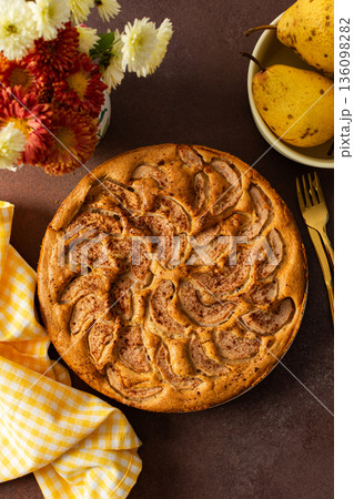 A fresh pear pie is placed on a cloth on a table. Nearby, a bowl holds ripe pears and a small vase has flowers. The setting looks warm and inviting for sharing a treat A fresh pear pie is placed on a cloth on a table. Nearby, a bowl holds ripe pears and a small vase has flowers. The setting looks warm and inviting for sharing a treat 136098282