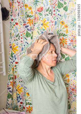 A 50-year-old woman stands in a vibrant bathroom, wrapping a towel around her head as she prepares for her morning routine. Flowers decorate the background, adding a lively touch A 50-year-old woman stands in a vibrant bathroom, wrapping a towel around her head as she prepares for her morning routine. Flowers decorate the background, adding a lively touch 136098305