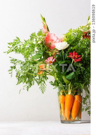 A bouquet of roses, gladioli, zinnias, and eustomas stands in water. Carrots with green tops are part of the arrangement on a plain table against a light background 136098619