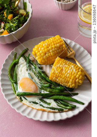 A healthy breakfast is served on a pink table. There are plates with asparagus, a fried egg, corn, and a fresh salad. Grapes and sunflower seeds are on the side A healthy breakfast is served on a pink table. There are plates with asparagus, a fried egg, corn, and a fresh salad. Grapes and sunflower seeds are on the side 136098647