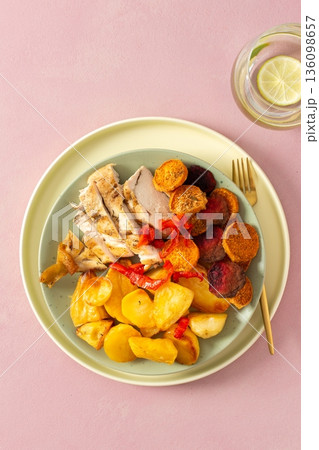 A plate holds baked potato, sliced baked chicken breast, and roasted vegetables like carrots, peppers, and beets. A drink sits nearby with a slice of lime A plate holds baked potato, sliced baked chicken breast, and roasted vegetables like carrots, peppers, and beets. A drink sits nearby with a slice of lime 136098657