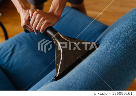 Close-up cropped shot of unrecognizable cleaning service worker using wet vacuum cleaner to remove dirt and stains from blue sofa, ensuring hygienic cleaning process. Concept of housekeeping Close-up cropped shot of unrecognizable cleaning service worker using wet vacuum cleaner to remove dirt and stains from blue sofa, ensuring hygienic cleaning process. Concept of housekeeping 136099161