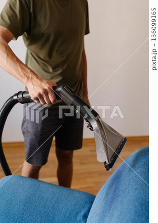 Vertical cropped shot of young man using washing vacuum cleaner to remove dirt and stains from blue sofa in home or apartment, providing efficient and thorough upholstery cleaning, close-up. Vertical cropped shot of young man using washing vacuum cleaner to remove dirt and stains from blue sofa in home or apartment, providing efficient and thorough upholstery cleaning, close-up. 136099165