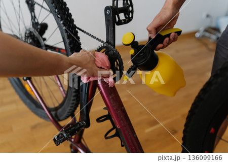 Close-up of two MTB biker male cleaning mountain bike using water sprayer and cloth, removing dirt and grime from frame at workshop, performing detailed maintenance after trail riding. 136099166