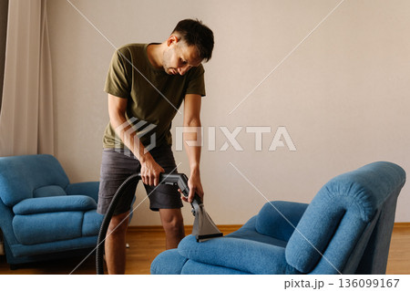 Young man in causal clothing using washing vacuum cleaner to clean blue sofa in home setting, demonstrating modern approach to household chores and upholstery maintenance. 136099167