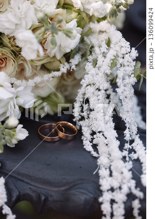 Gold wedding rings beside white floral bouquet on dark metal bench Gold wedding rings beside white floral bouquet on dark metal bench 136099424