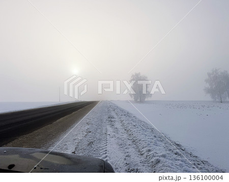 Asphalt road with a snow-covered shoulder recedes into the distance. The sky is in a frosty haze, through which the solar disc is visible. A car hood is in the foreground. Winter landscape. 136100004