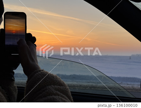 A driver of a passenger car has slightly opened the car window and is filming the sunset with a smartphone. A snow-covered field is visible to the horizon. Above it, a red solar disc and the glow of t 136100010