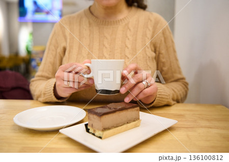 Woman In Beige Knit Sweater Sips Coffee Beside Chocolate Layer Cake Dessert At Table 136100812