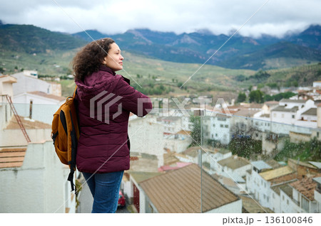 Woman With Brown Jacket And Backpack Overlooking A Mountain Town From Glass Balcony 136100846