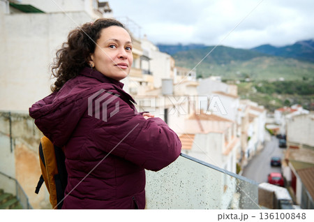 Thoughtful Woman Standing On Balcony In Burgundy Jacket Overlooking Scenic Town And Hills 136100848