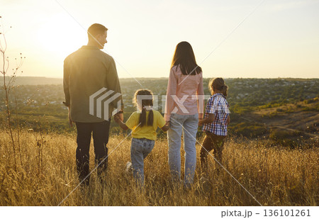 Family holding hands in golden meadow at sunset, looking over scenic valley 136101261