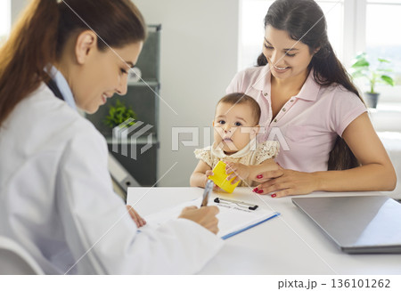 Doctor recording notes as mother holds baby during gentle pediatric consultation Doctor recording notes as mother holds baby during gentle pediatric consultation 136101262