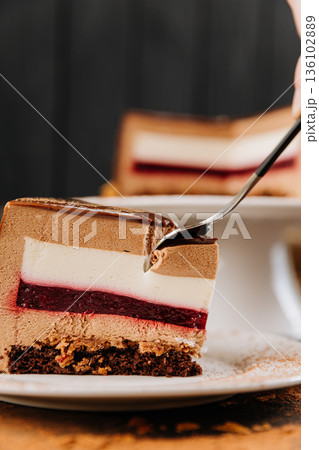 Fork reaching for layered chocolate mousse cake slice with berry jelly on white plate Fork reaching for layered chocolate mousse cake slice with berry jelly on white plate 136102889