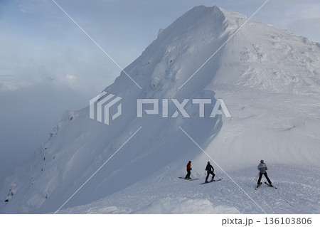Three skiers stopped on a ridge and look towards a massive snow-covered slope Three skiers stopped on a ridge and look towards a massive snow-covered slope 136103806
