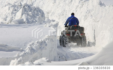 A person rides an ATV through a narrow passage carved between massive snowbanks 136103810
