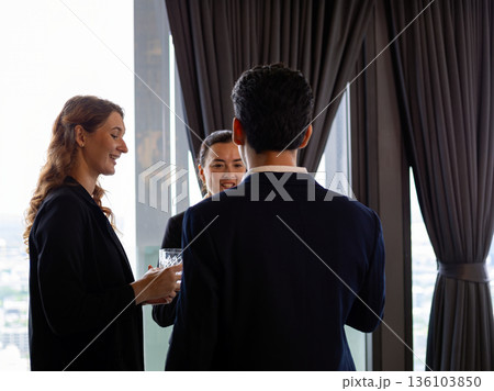 Business professionals networking and talking together in a modern office with city view, wearing formal suits, representing corporate communication, teamwork, partnership, and professional 136103850