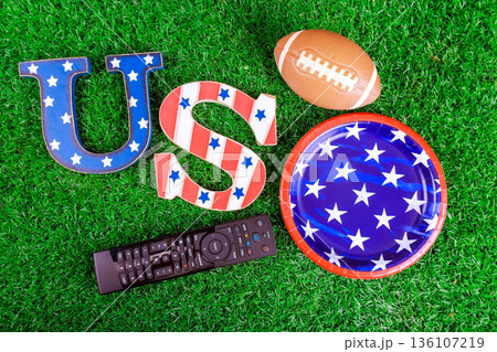 Decorations items for sports event include American football, remote, themed plates on grassy surface. 136107219