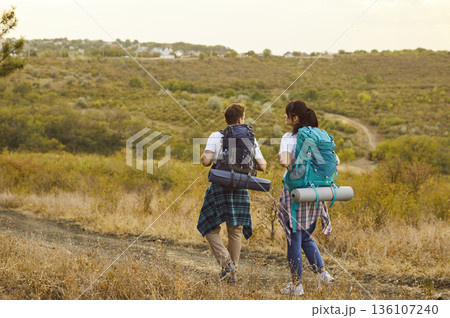 Hikers couple with backpacks hiking on path through field and meadow, back view 136107240