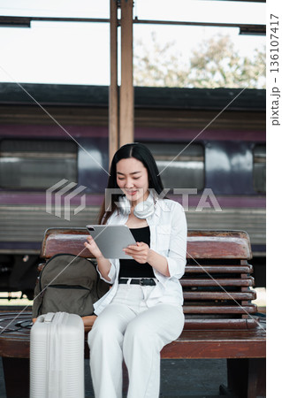 Woman sitting on a bench at a train station with a tablet and luggage, ready for travel. 136107417