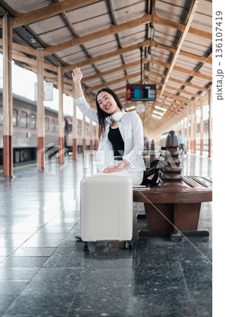 Smiling traveler at a train station with luggage, waving and ready for a journey. Modern travel and exploration concept. Smiling traveler at a train station with luggage, waving and ready for a journey. Modern travel and exploration concept. 136107419