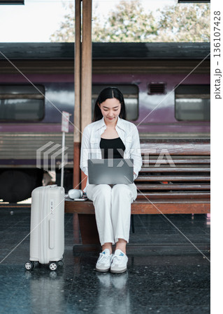 Woman in casual attire using a laptop at a train station, showcasing modern travel and remote work. 136107428