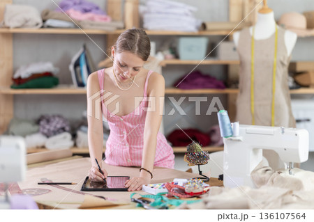 Young female dressmaker drawing in tablet in sewing workshop 136107564