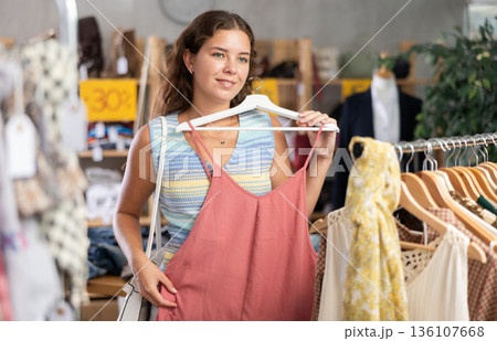 Positive young woman choosing something for summer in clothing store 136107668
