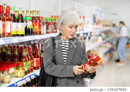 Senior woman customer is thinking about buying spicy and unusual red chilli tsuyu sauce. 136107683