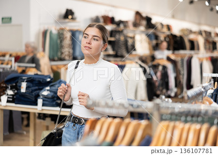 Positive joyful young woman looking for fashionable clothes in clothing boutique Positive joyful young woman looking for fashionable clothes in clothing boutique 136107781