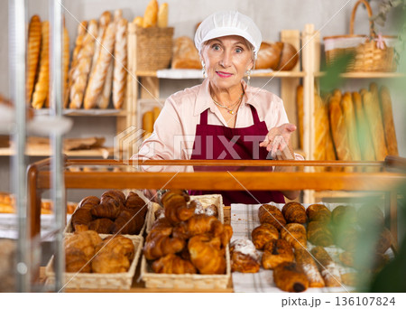 Portrait of elderly woman baker standing behind display case with fresh croissants. Offering to buy fresh baked goods Portrait of elderly woman baker standing behind display case with fresh croissants. Offering to buy fresh baked goods 136107824