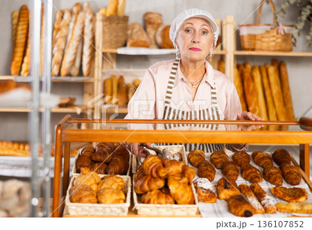 Senior woman employee puts croissants in window, arranges display of goods at bakery. 136107852