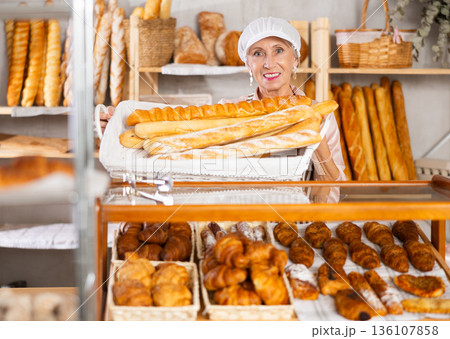 Senior female baker worker sells baguette, shows many products Senior female baker worker sells baguette, shows many products 136107858