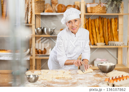 Experienced elderly woman baker using knife to cut raw dough to make croissants 136107862