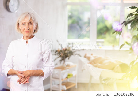 Female elderly physiotherapist poses against background of spa salons office. 136108296