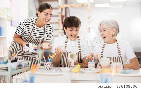 Cheerful older woman with young adult children painting pottery in family workshop 136108306