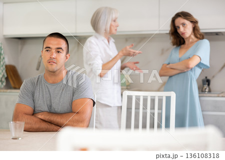 Elderly woman quarrels with couple in kitchen Elderly woman quarrels with couple in kitchen 136108318