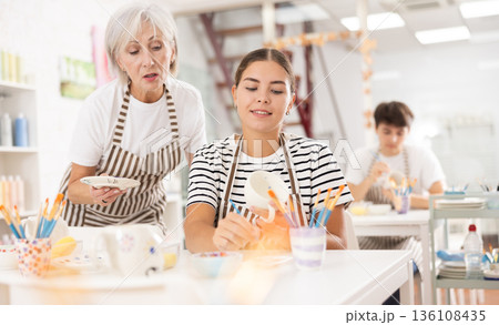 Elderly mother guiding adult daughter painting mugs in family pottery workshop Elderly mother guiding adult daughter painting mugs in family pottery workshop 136108435