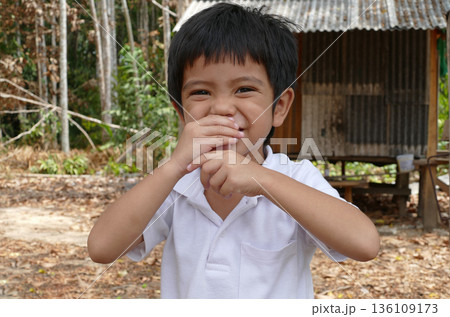 Asian Thai little boy use hands close his mouth during smiling and standing front of old small wood house in garden 136109173