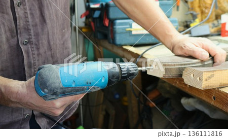 Close-up of carpenter's hands with blue screwdriver and wooden curved cut piece or furniture fragment on work surface while drilling holes on end face of piece in workshop Close-up of carpenter's hands with blue screwdriver and wooden curved cut piece or furniture fragment on work surface while drilling holes on end face of piece in workshop 136111816