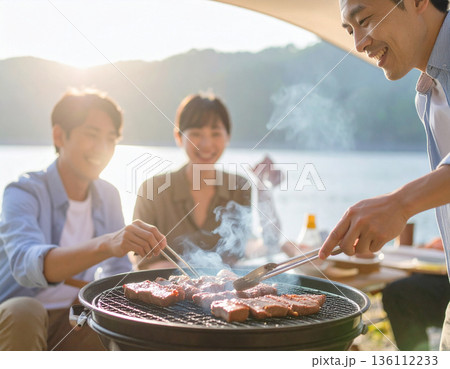 水辺でキャンプ、バーベキュー料理を楽しむ家族 136112233