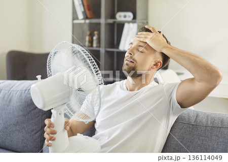 Exhausted man cooling himself with fan sit on couch in living room Exhausted man cooling himself with fan sit on couch in living room 136114049