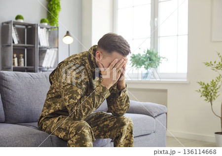 Young man in military camouflage uniform sits on couch expressing deep sadness or stress 136114668