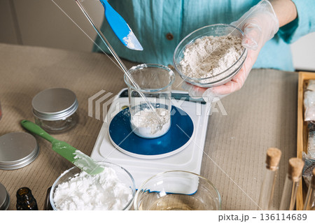 Hands use blue spatula to transfer white powder from bowl into glass beaker on digital scale. lab-style skincare aesthetic, cosmetic formulation process, mindful chemistry, apothecary workspace. 136114689